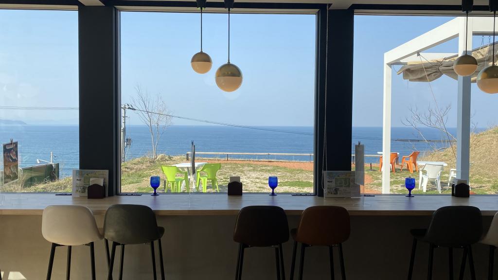 View of the ocean from inside a cafe. There are plastic chairs and tables outside.